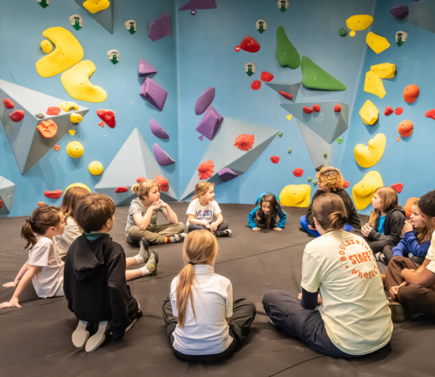 A group of children and two instructors sit in a circle on mats in a Brooklyn indoor climbing gym, colorful climbing holds covering the blue walls behind them—a perfect setting for Summer Adventures.