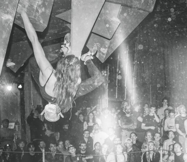 A climber hangs from an overhanging indoor climbing wall during Boulderfest 2025 in Brooklyn, surrounded by a crowd of spectators watching intently.