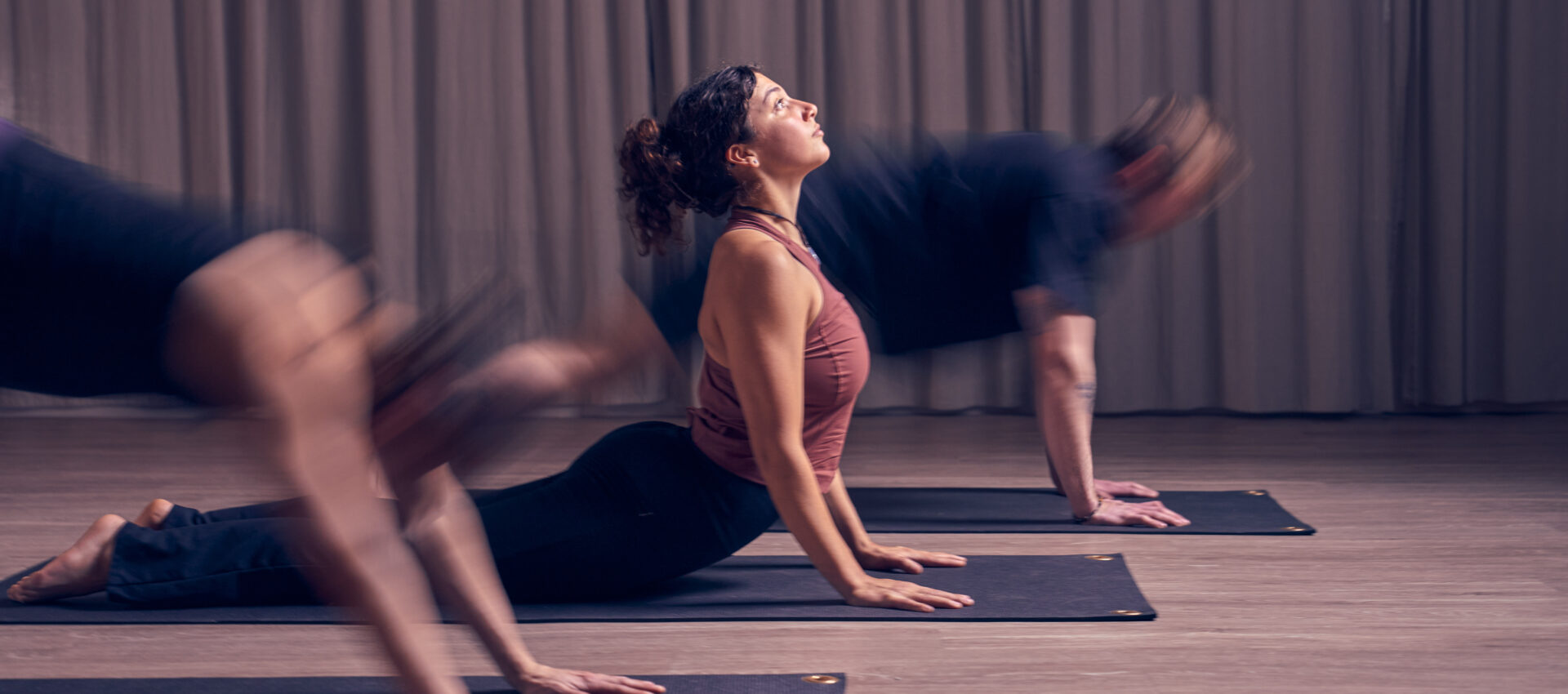 Three people practice yoga indoors on mats, performing the upward-facing dog pose. The central woman is in focus, with blurred motion on the sides and a curtain in the background—perfect for your Yoga Class Calendar inspiration.