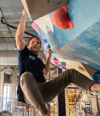 A man in a black shirt and tan pants is climbing an indoor rock wall, gripping colorful holds and looking focused. The modern climbing gym features industrial elements, creating the perfect environment for this challenging climb.