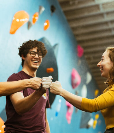 Three people smile and give each other a fist bump in front of a blue indoor climbing wall with colorful holds at a climbing gym in Seattles University District.