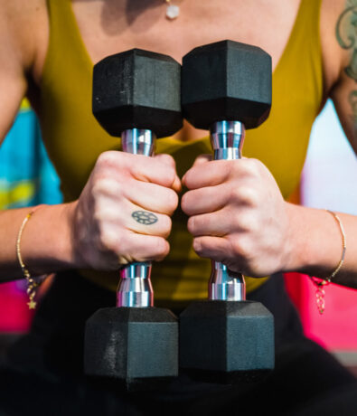 A person in a yellow tank top grips two black hexagonal dumbbells, holding them together vertically at an indoor rock climbing gym. They have a small tattoo on one finger and wear delicate bracelets on both wrists.