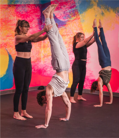 Four people are in a colorful studio at an Austin climbing gym; one assists another with a handstand in the foreground, while two others practice handstands independently in the background.