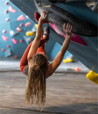 A woman with long braided hair, wearing an orange outfit, climbs an indoor bouldering wall, gripping large black holds while suspended horizontally above padded flooring.