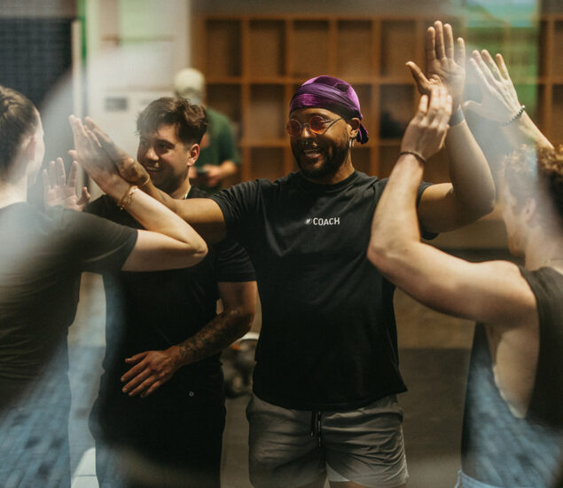 A group of people, including a smiling coach wearing a purple headscarf and black COACH shirt, stand in a circle indoors, giving high-fives—celebrating teamwork and the Membership Benefits at Austin Bouldering Project.