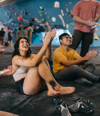A woman and a man sit on the floor of an indoor rock climbing gym, smiling and clapping. Climbing shoes rest nearby, with colorful holds dotting the wall in the background.