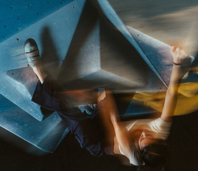A person with long hair climbs an indoor bouldering wall with large blue holds and yellow grips, reaching upward with one arm. The motion blur suggests dynamic movement at a Salt Lake City gym offering 24/7 access.