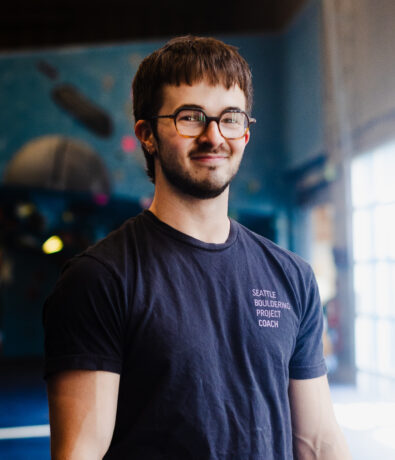 A man with short brown hair and glasses, wearing a dark Seattle Bouldering Project Coach T-shirt, stands indoors with a slight smile. The background is softly blurred with blue wall and climbing gym elements visible.