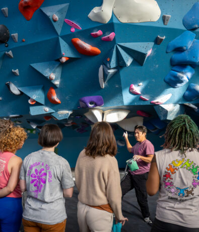 A group of people stands in front of an instructor under a large indoor climbing wall at Brooklyn Climbing Yoga and Fitness. Colorful holds and geometric volumes surround them as the instructor gestures, preparing the group for a climbing session.