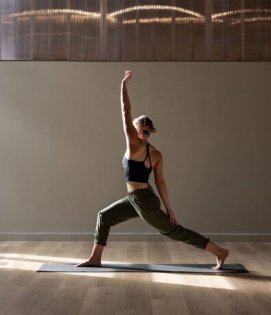A woman practices yoga indoors in Fremont, performing a warrior pose on a mat. She wears a black sports bra and olive-green pants, with one arm extended upward and sunlight streaming across the upper walls through a window.