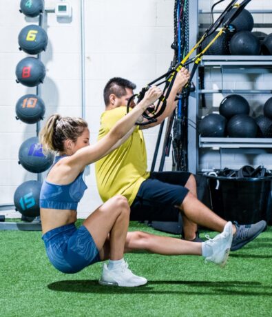 Two people exercise indoors in the Design District using TRX straps, performing single-leg squats on artificial turf. Weighted balls and gym equipment can be seen in the background.