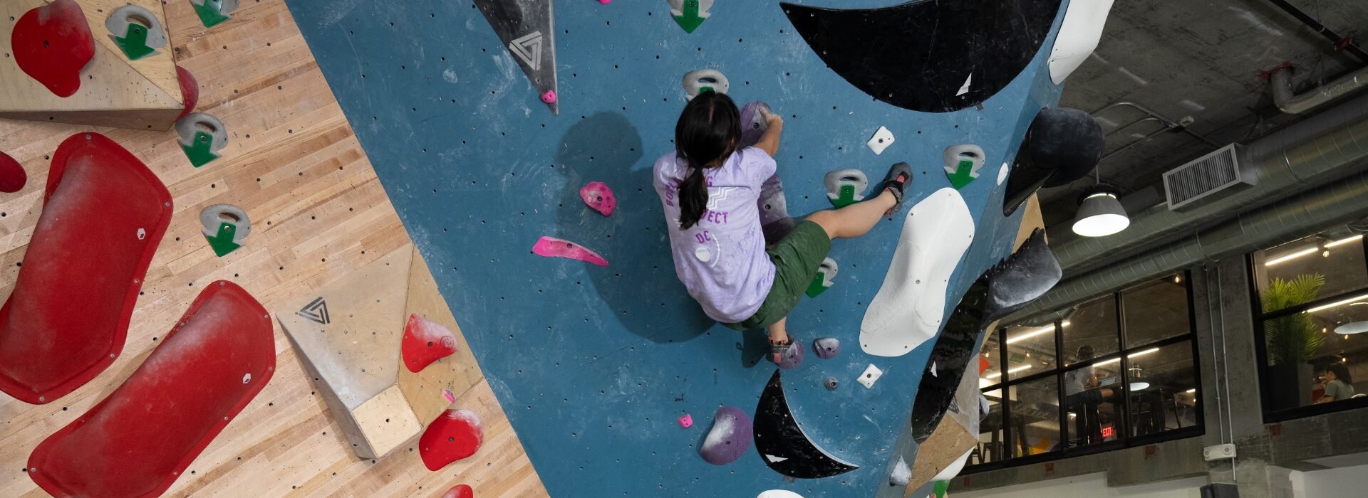 A person with a ponytail is climbing an indoor bouldering wall with colorful holds, surrounded by wooden panels and large windows in a modern gym.