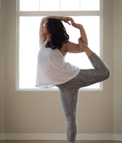 A woman practices yoga indoors in Fremont, standing on one leg in a dancer pose near a large bright window, her silhouette highlighted by the sunlit upper walls, wearing a white tank top and patterned leggings.