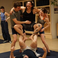 Four people at Brooklyn Climbing Yoga and Fitness are laughing and balancing together in a playful acro-yoga pose indoors, while another person smiles in the background, enjoying the energetic and supportive atmosphere.