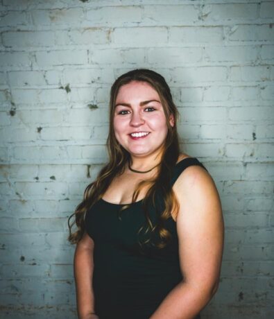 A young woman with long, wavy brown hair wearing a sleeveless black dress smiles while standing in front of a light-colored brick wall.