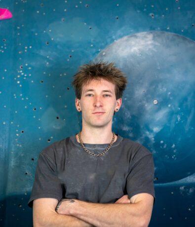 A young man with short, spiky hair and earrings stands with arms crossed in front of a blue wall painted with a large, grayish planet and small stars—his confident look hinting at countless hours spent on his latest bouldering project.