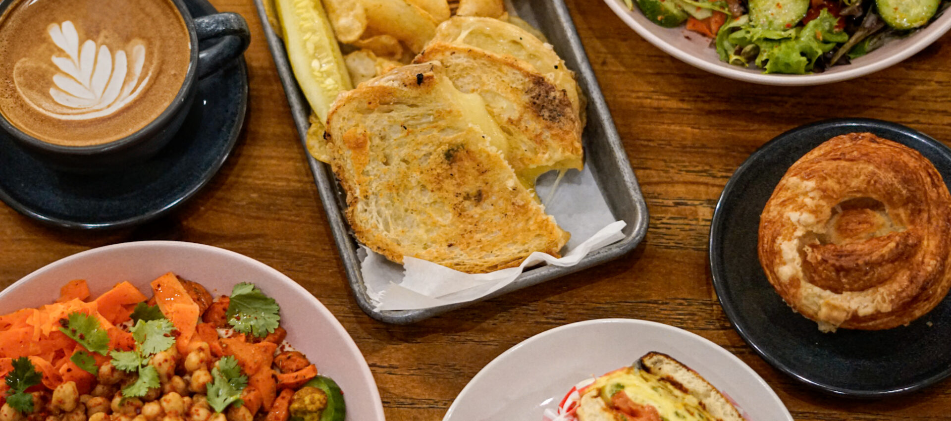 A wooden table at West Wall Cafe set with a cup of latte art coffee, a grilled cheese sandwich with chips and a pickle, a fresh salad, a chickpea and vegetable bowl, a croissant, and a sandwich with tomato and lettuce.