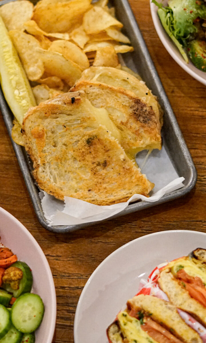 A wooden table at West Wall Cafe set with a cup of latte art coffee, a grilled cheese sandwich with chips and a pickle, a fresh salad, a chickpea and vegetable bowl, a croissant, and a sandwich with tomato and lettuce.