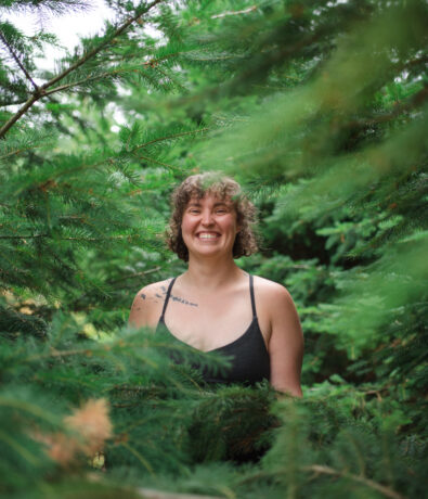 A smiling person with curly hair and a sleeveless black top stands among lush green pine trees on the Upper Walls in Fremont, partially hidden by the branches.