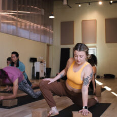 Several people practice yoga indoors on mats, using cork yoga blocks for support. The focus is on a woman in a yellow top and brown pants in a low lunge position, with natural light filling the studio.