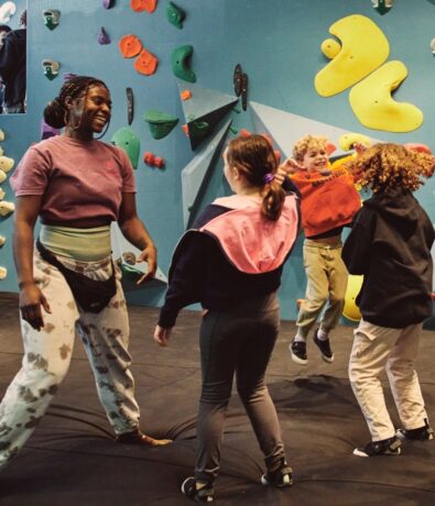 A woman and three children laugh and play together at an indoor climbing gym in Brooklyn, standing near colorful climbing holds. The lively scene captures the joy of Summer Adventures 2024, with one child mid-jump.