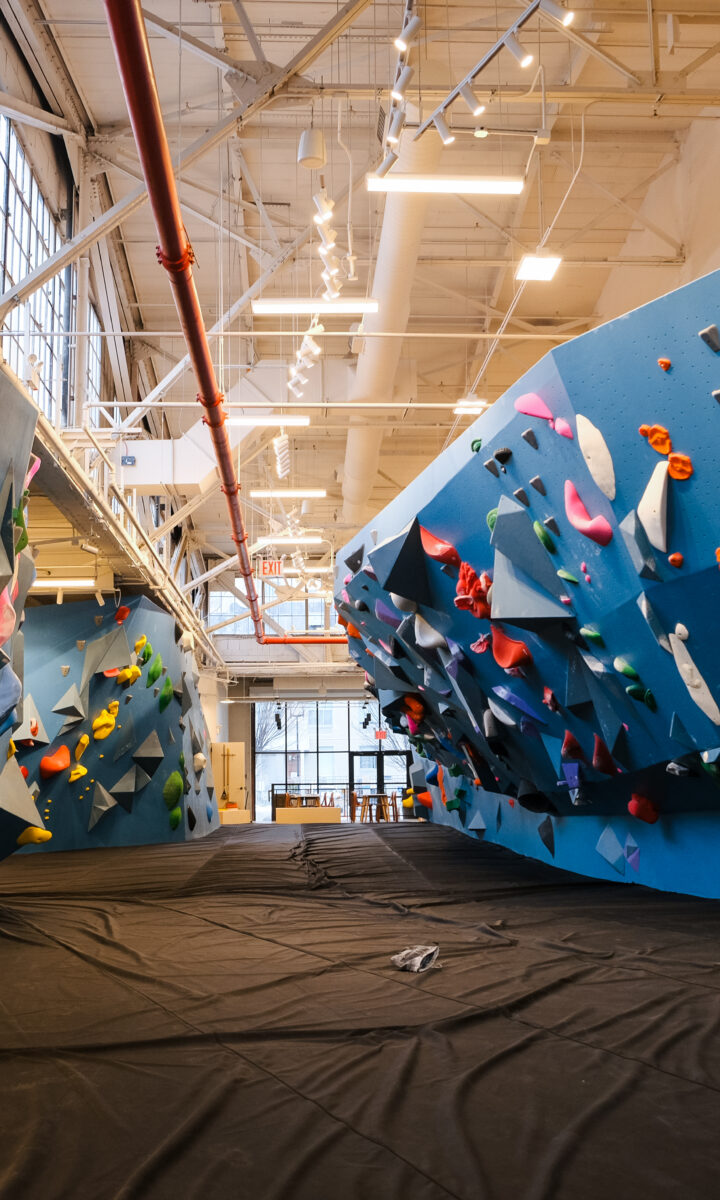 Brooklyn Climbing Yoga and Fitness offers an indoor rock climbing gym with large blue walls, colorful holds, padded black flooring, and bright lighting. Exposed ceiling beams and windows on one wall create an open, inviting space. No people are present.