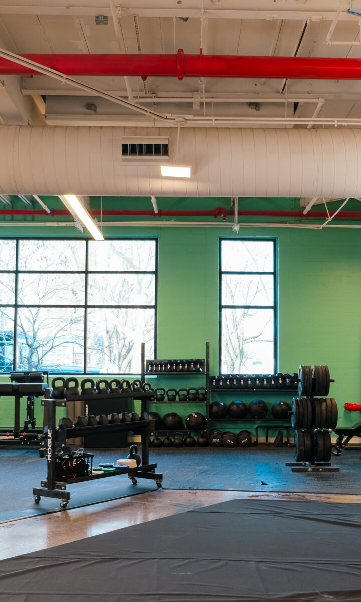 A brightly lit gym with large windows, green and brick walls, weight racks, benches, and neatly arranged exercise equipment awaits at Brooklyn Climbing Yoga and Fitness.