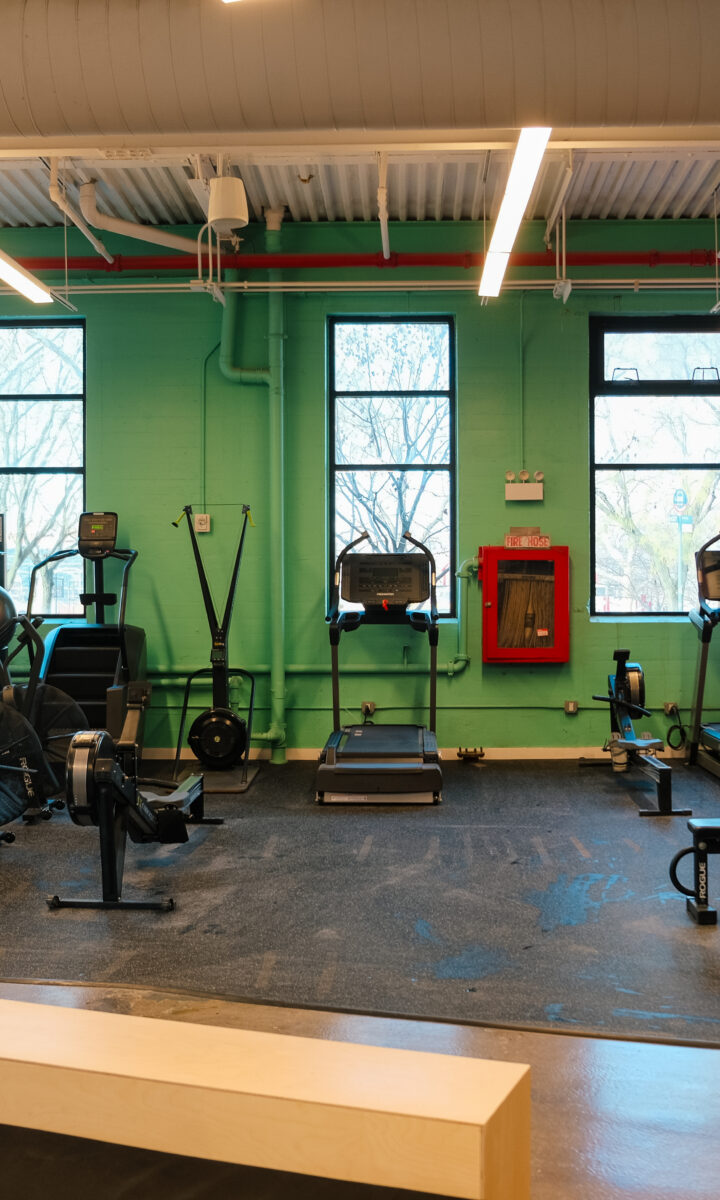 A modern gym at Brooklyn Climbing Yoga and Fitness with cardio machines, stationary bikes, weightlifting equipment, and a colorful mural. Large windows fill the clean, organized space with natural light.
