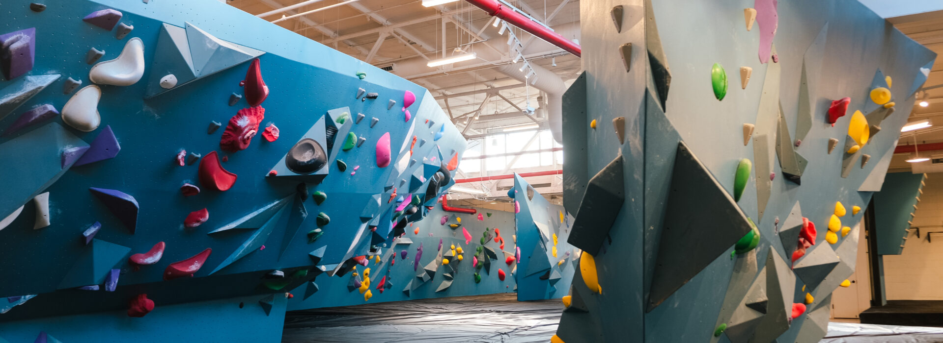 Indoor rock climbing gym with blue climbing walls, colorful holds, and black padded flooring beneath. Brooklyn Climbing Yoga and Fitness offers a well-lit space with overhead lights and multiple climbing routes of varying difficulty.