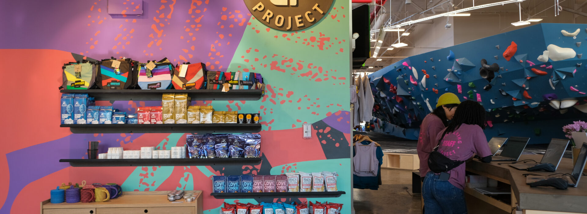 A colorful bouldering gym reception area with snacks and gear on display. Two people stand at the counter, while climbing walls with holds and climbers are visible in the background, highlighting Brooklyn Climbing Yoga and Fitness vibes.