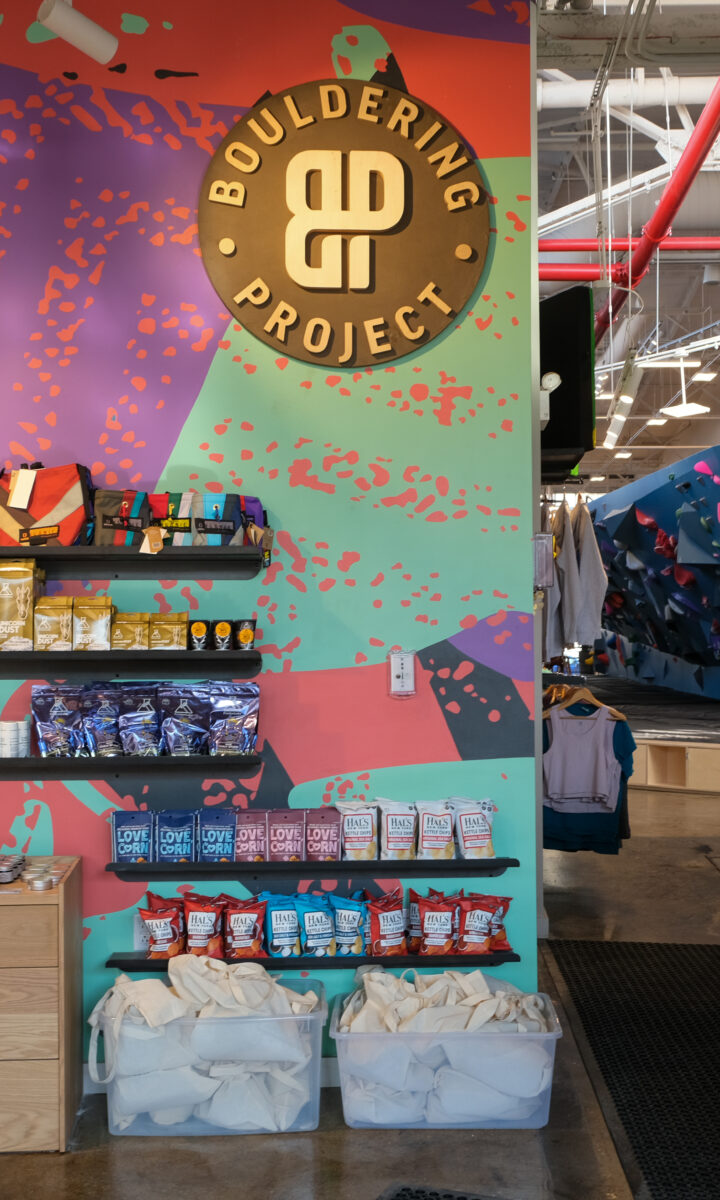 A colorful bouldering gym reception area with snacks and gear on display. Two people stand at the counter, while climbing walls with holds and climbers are visible in the background, highlighting Brooklyn Climbing Yoga and Fitness vibes.