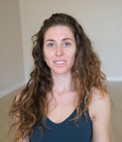 A woman with long, curly brown hair and blue eyes, wearing a dark tank top, sits indoors against a plain light-colored wall—smiling softly after a Brooklyn Climbing Yoga and Fitness class.