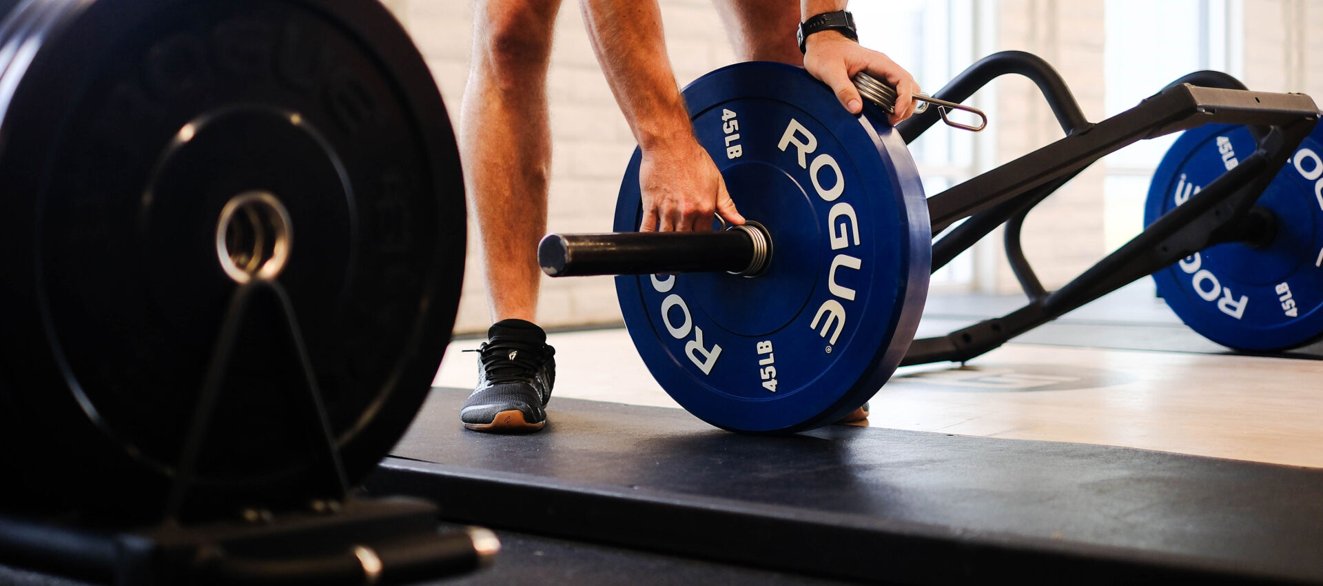 A person in Tempe adjusts blue Rogue weight plates on a barbell, preparing for weightlifting. Gym equipment and mats are visible in the background, setting the scene for personal training sessions.