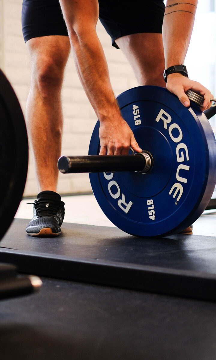 A person in Tempe adjusts blue Rogue weight plates on a barbell, preparing for weightlifting. Gym equipment and mats are visible in the background, setting the scene for personal training sessions.