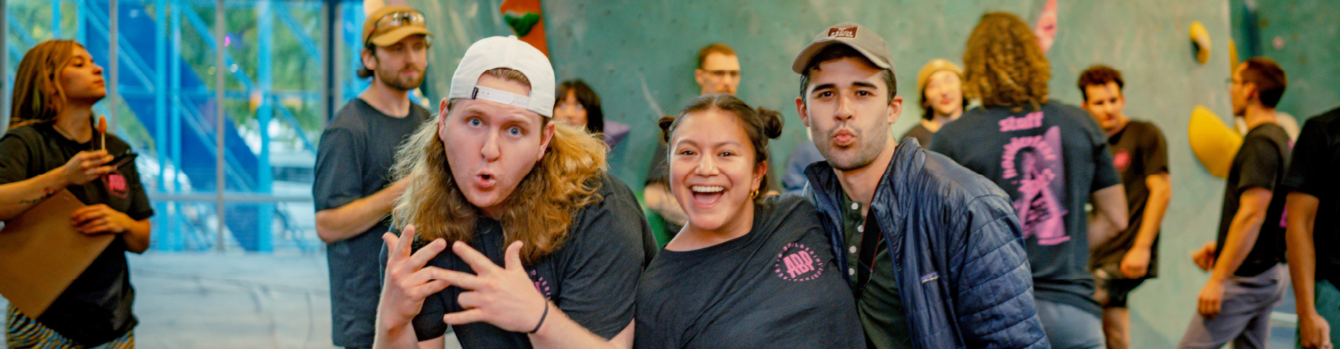A group of people pose and smile for the camera in a lively indoor setting, with some making playful expressions and gestures. Others talk and laugh in the background, creating a cheerful, social atmosphere.