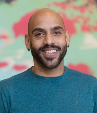 A smiling man with a shaved head, beard, and hoop earrings, wearing a teal shirt, stands in front of a colorful blurred background—ready to offer personal training in Minneapolis.