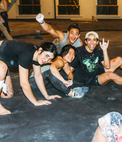 Four young adults pose playfully on a gym floor at Climbing Tempe, covered in chalk dust, smiling and making energetic hand gestures, with a casual, fun atmosphere in this vibrant indoor climbing gym.
