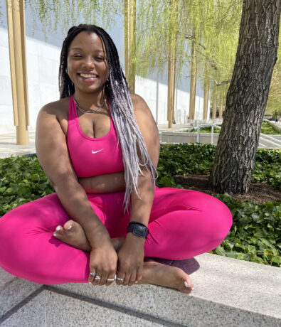 A woman in a bright pink athletic outfit sits cross-legged on a stone ledge outdoors, smiling at the camera. She has long braided hair, a smartwatch, and is surrounded by greenery and trees.