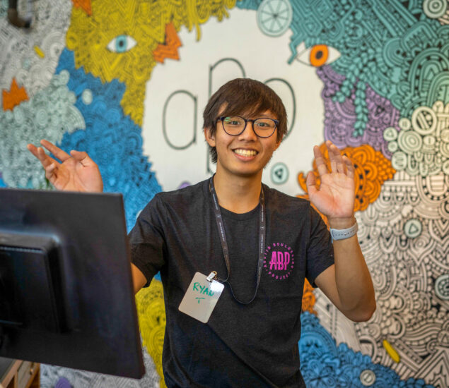 A smiling person wearing glasses, a lanyard, and a name tag stands in front of a colorful, doodle-style mural, cheerfully raising both hands near their face. A computer monitor for managing Group Reservations is visible in the foreground.