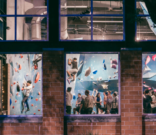 People are indoor rock climbing on colorful walls inside a large gym, viewed through windows from outside at night; several climbers are on the walls while others watch or wait below. Group reservations make this an ideal spot for friends to climb together.