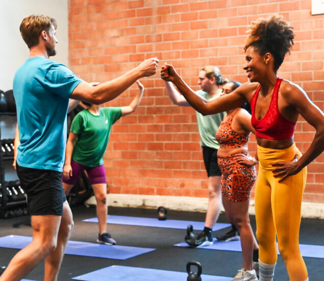 A group fitness class with four people smiling and fist bumping. They are wearing colorful workout clothes and standing on exercise mats in front of a brick wall, with kettlebells on the floor nearby—perfect for Group Reservations.