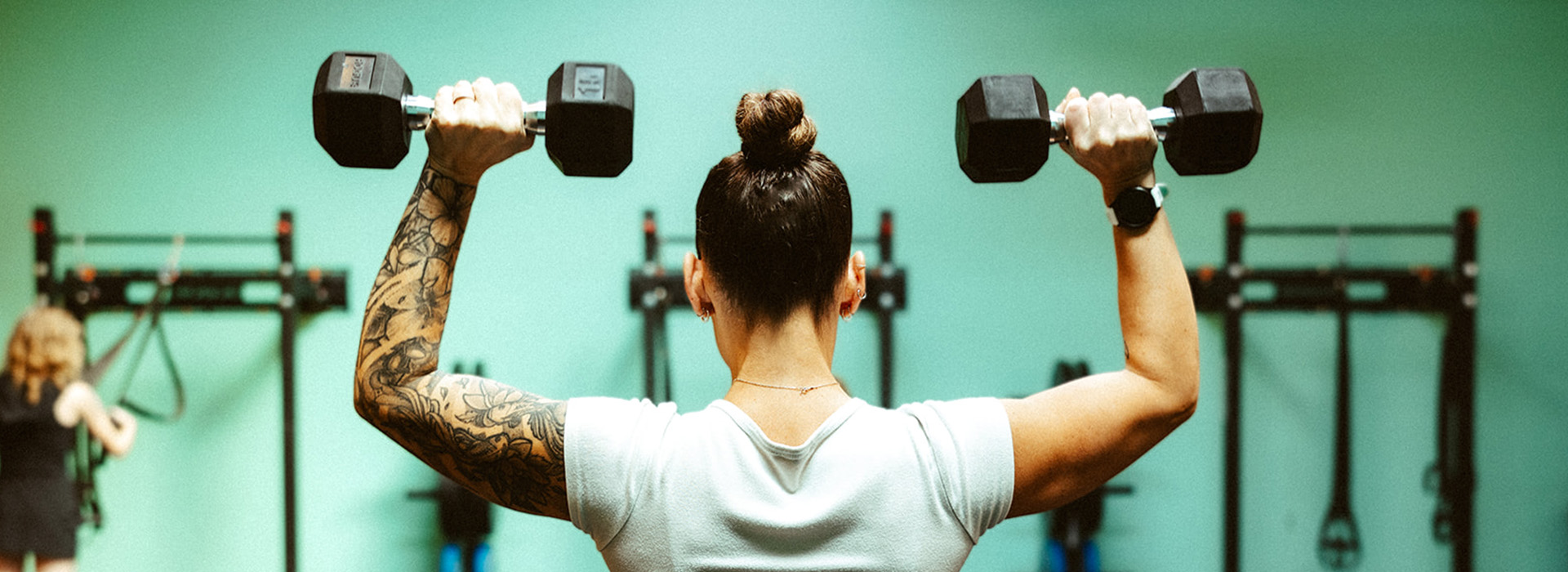 A woman with a tattooed arm lifts two dumbbells overhead in a gym, seen from behind. Gym equipment and another person are visible in the background.