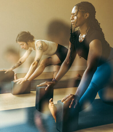 Two women practice yoga indoors at a climbing gym in the University District, Seattle, using yoga blocks for support in a stretching pose. Both appear focused, wearing athletic wear as natural light fills the room.