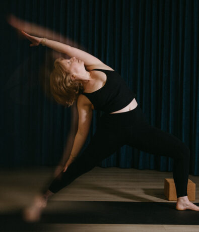 A person in black athletic wear practices a yoga pose indoors at a climbing gym in the University District, Seattle, stretching one arm overhead with blurred motion, while a yoga block and dark curtain appear in the background.