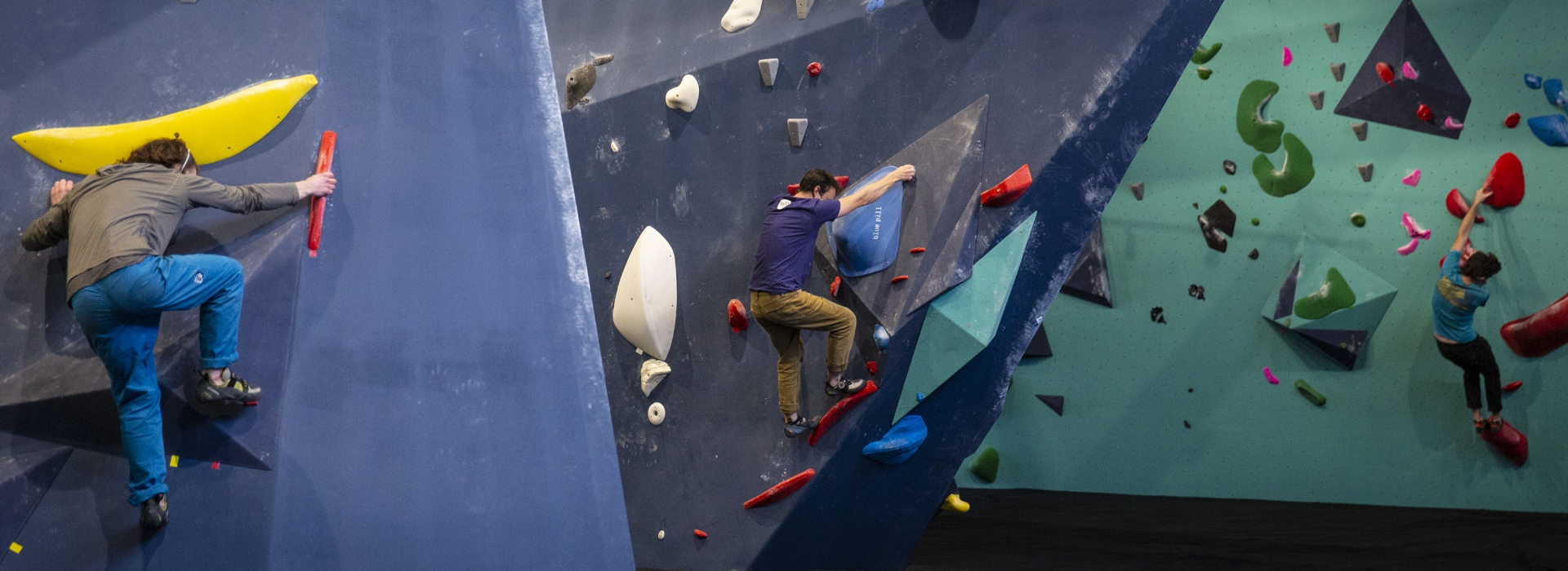 Three people are indoor rock climbing at Upper Walls in Fremont, tackling colorful, angled climbing walls with various shaped holds. Each climber follows a different route, using hands and feet to navigate the challenging surfaces.