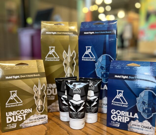 Boxes and tubes of FrictionLabs climbing chalk and liquid chalk, labeled Unicorn Dust, Gorilla Grip, and Secret Stuff, are displayed on a wooden surface in a retail setting, with a blurred colorful MBP background indoors.