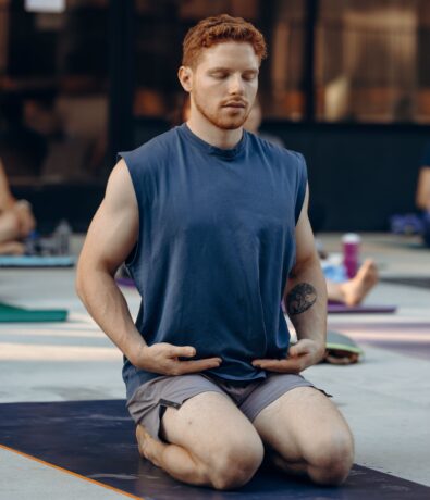 A man with red hair and a beard kneels on a yoga mat outdoors, eyes closed, hands resting on his thighs, practicing meditation or breathing exercises. His sleeveless blue shirt and gray shorts suggest he’s relaxing after climbing.