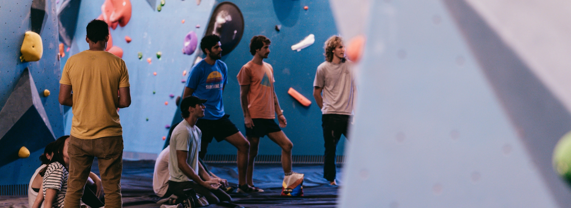 A group of young people, some standing and some sitting, gather inside a climbing gym with colorful holds on blue walls. They appear to be watching or discussing something—perfect for those interested in group reservations.