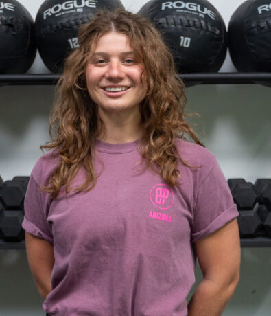 A young woman with wavy brown hair and a purple t-shirt stands and smiles in front of shelves with black medicine balls and dumbbells in a gym setting.