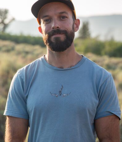 A bearded man in a blue t-shirt and black cap stands outdoors in a grassy field near SLC, with trees and mountains blurred in the background, sunlight softly illuminating the scene—an ideal setting for personal training sessions.